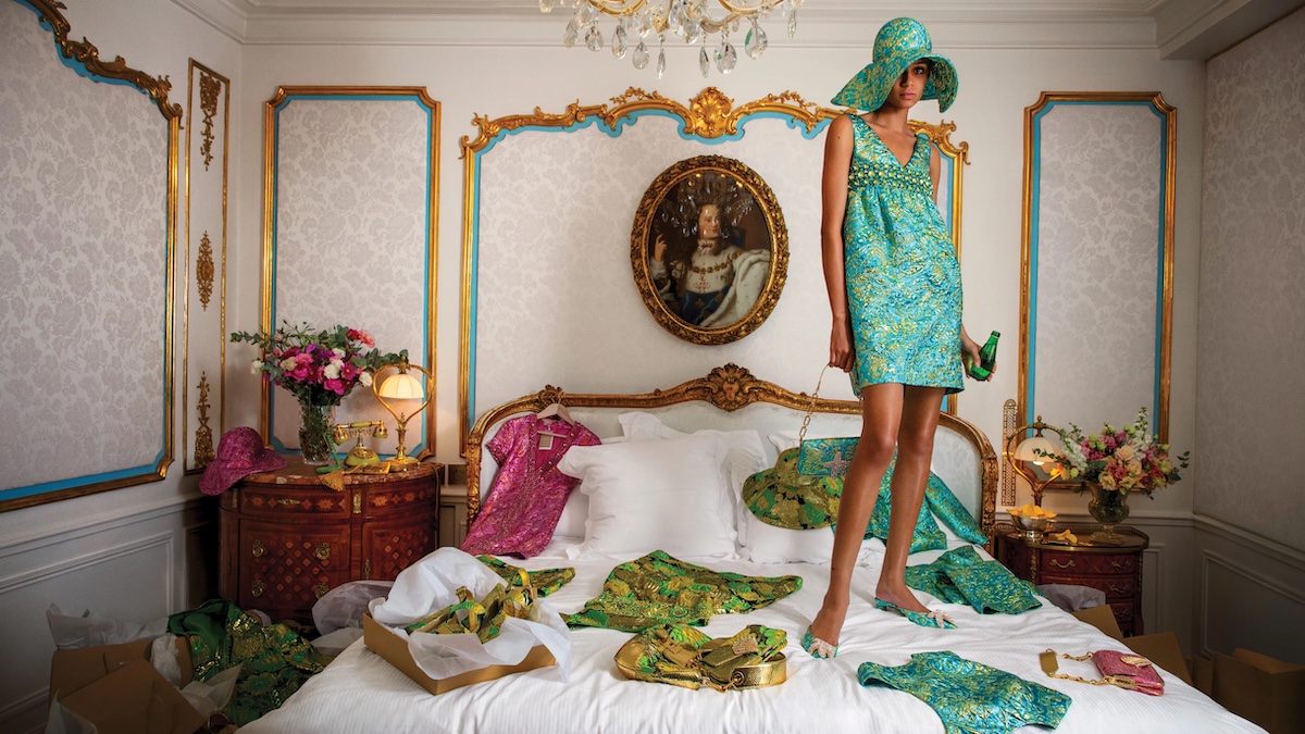 Woman in green and gold dress and matching shoes stands on bed with clothes strewn around her
